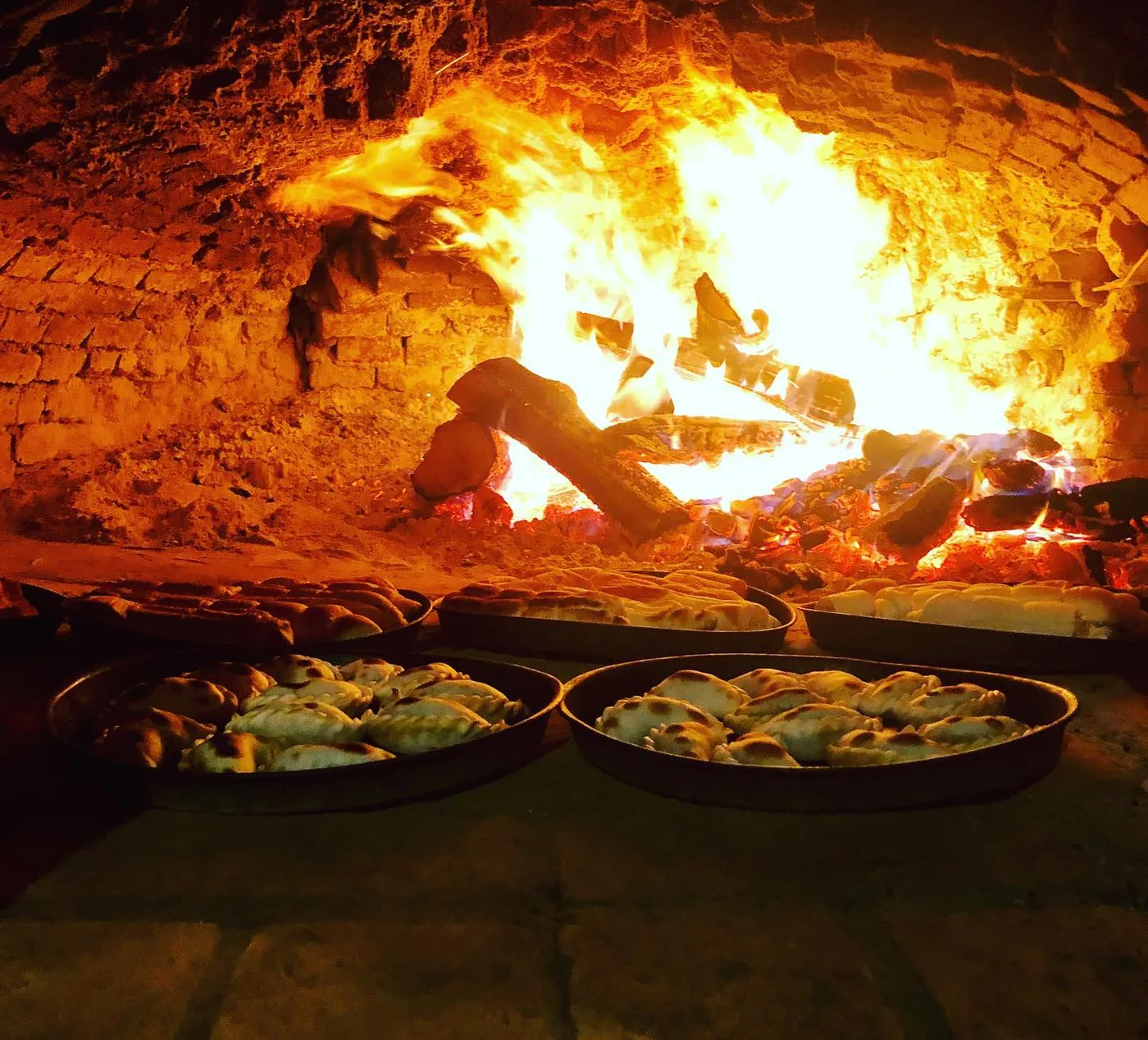 Horno de barro encendido a leña con bandejas de empanadas listas para hornear